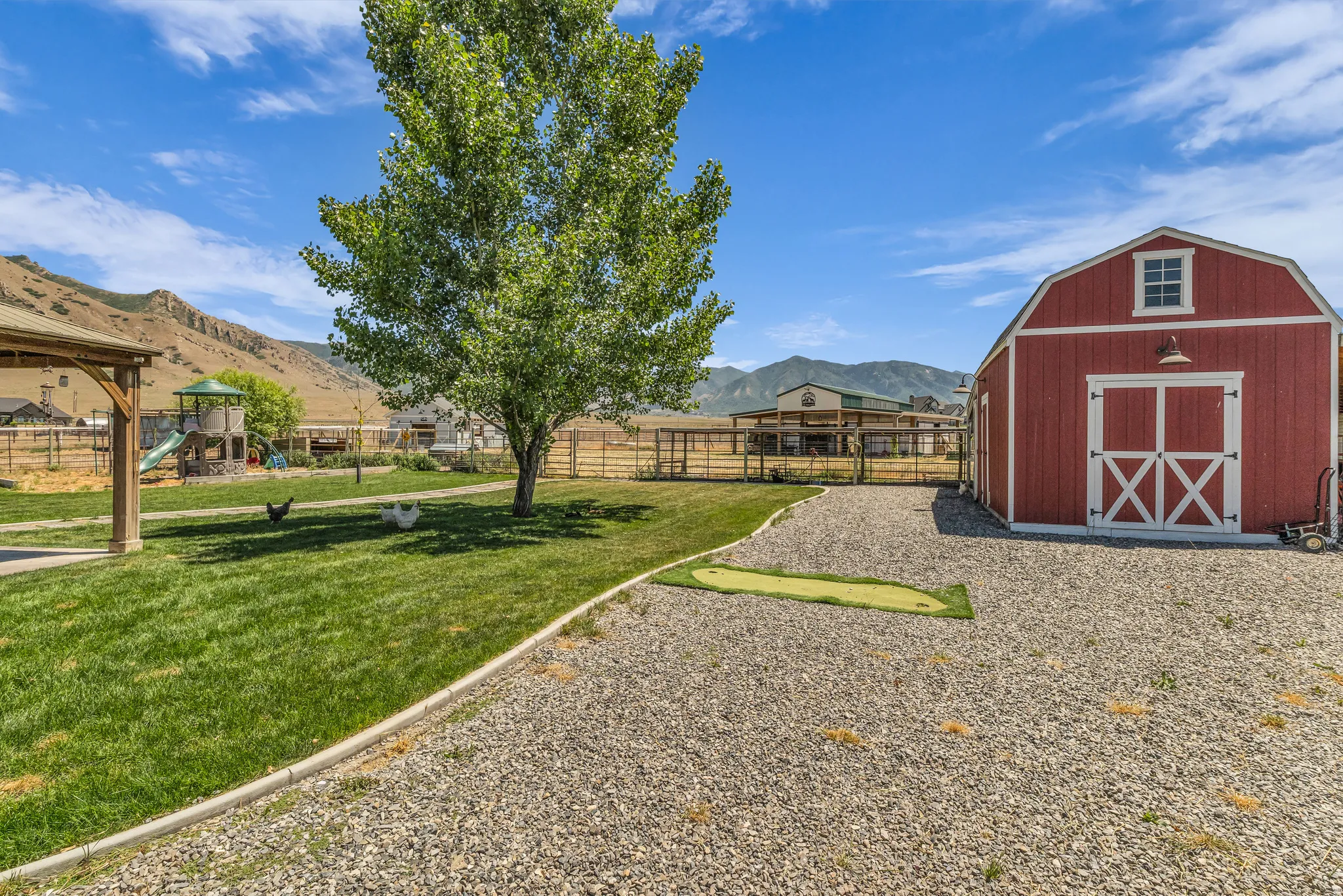 Expansive outdoor area featuring a red barn with white trim and barn doors, a gravel driveway, a manicured lawn with a mature shade tree, and a covered pergola
