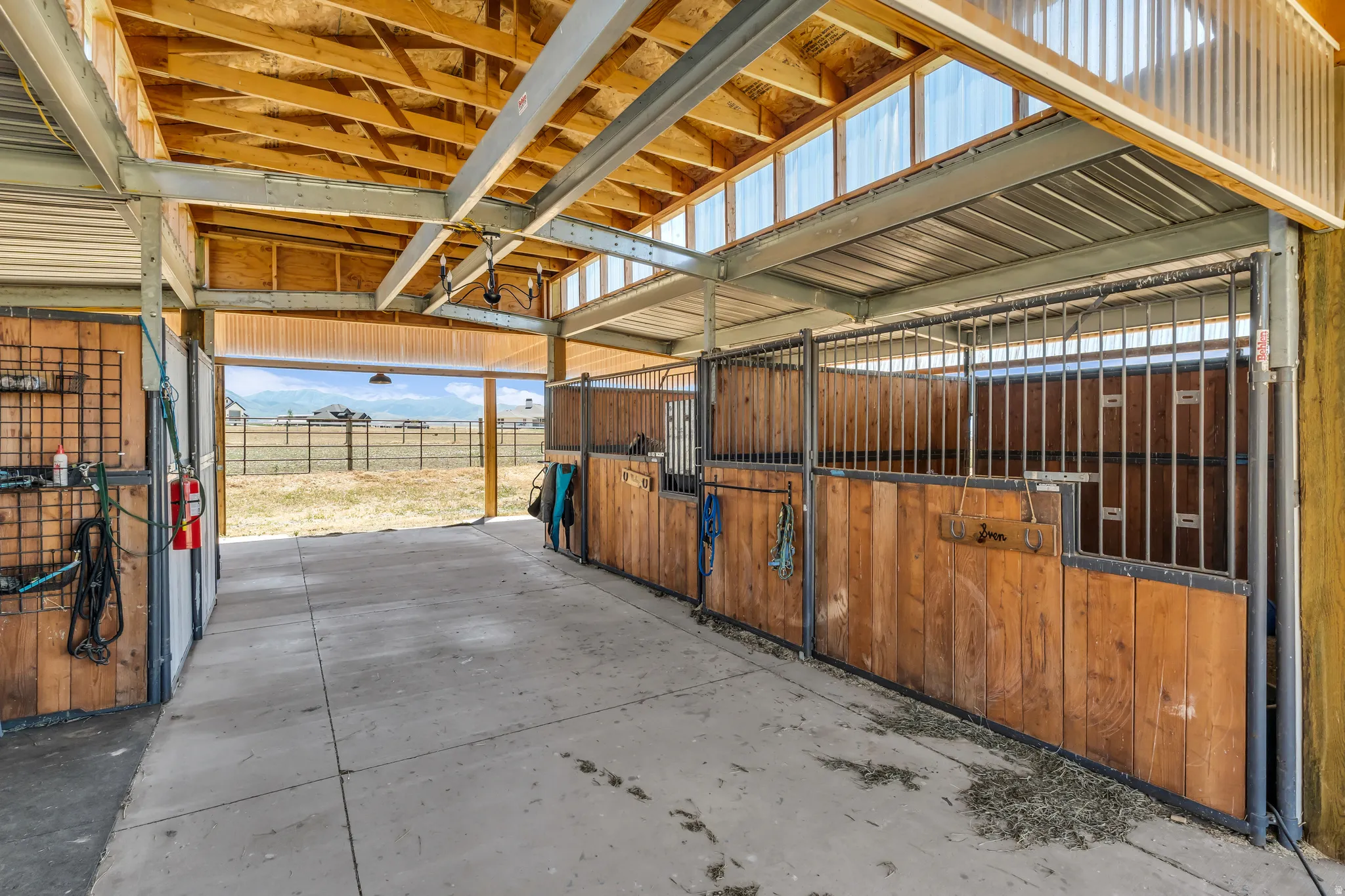 Barn interior featuring concrete flooring, multiple stalls with wood paneling and metal gating, an exposed wood beam ceiling, and translucent roof panels allowing for natural light