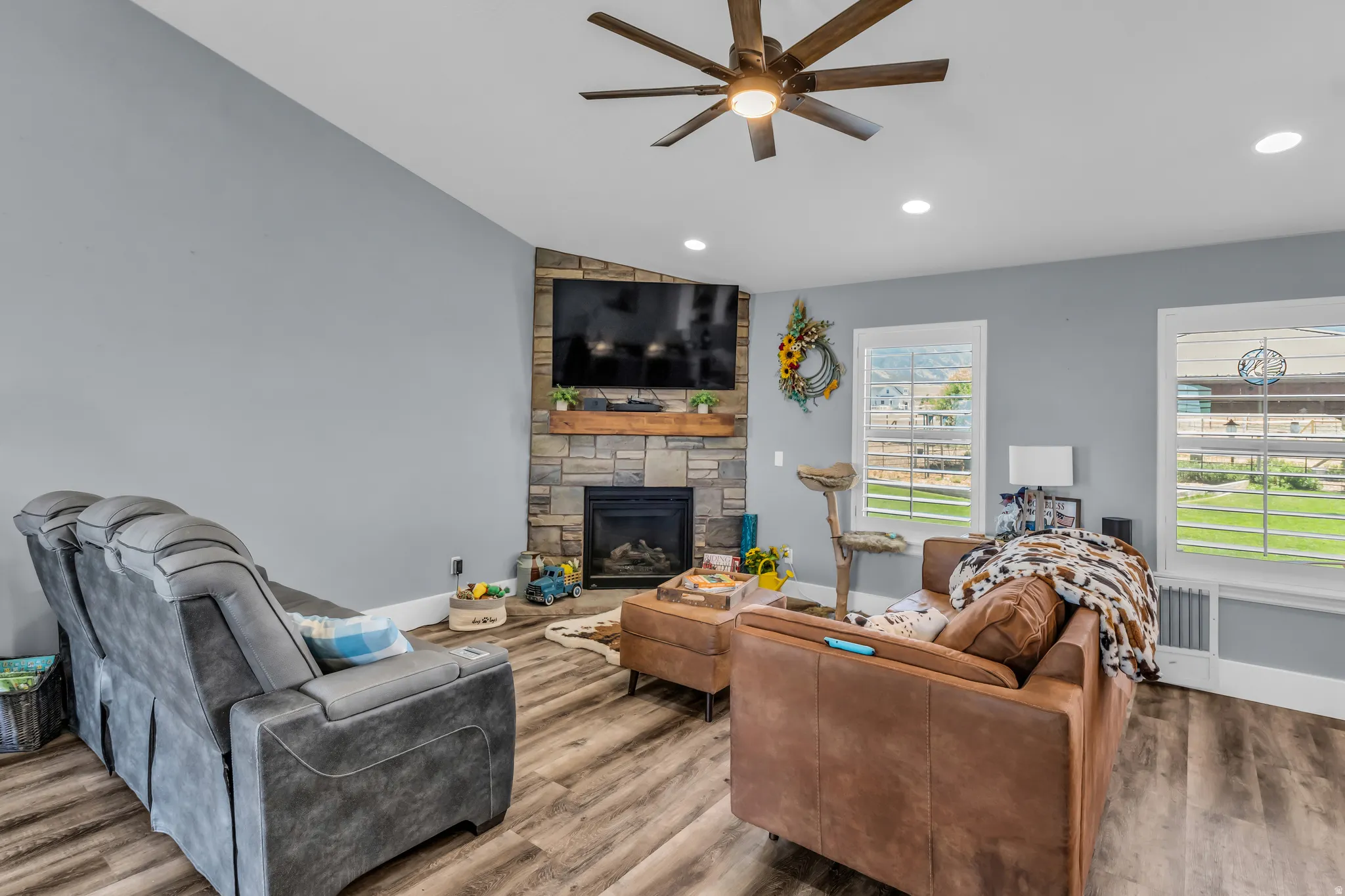 Living area featuring a stone fireplace with a wood mantle, wood-finish flooring, two windows, recessed lighting, and a ceiling fan