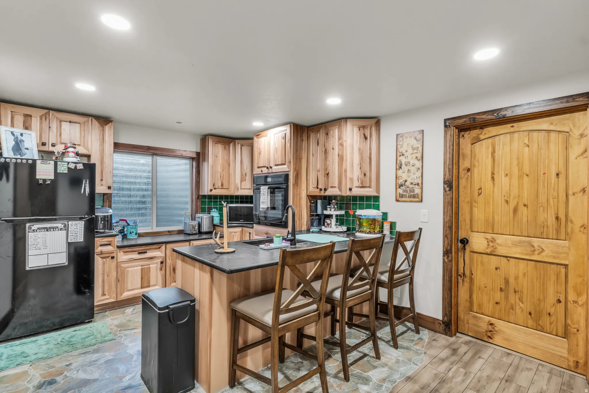 Kitchen featuring natural wood cabinetry with countertop seating, integrated black appliances, a green tile backsplash, and slate-style flooring