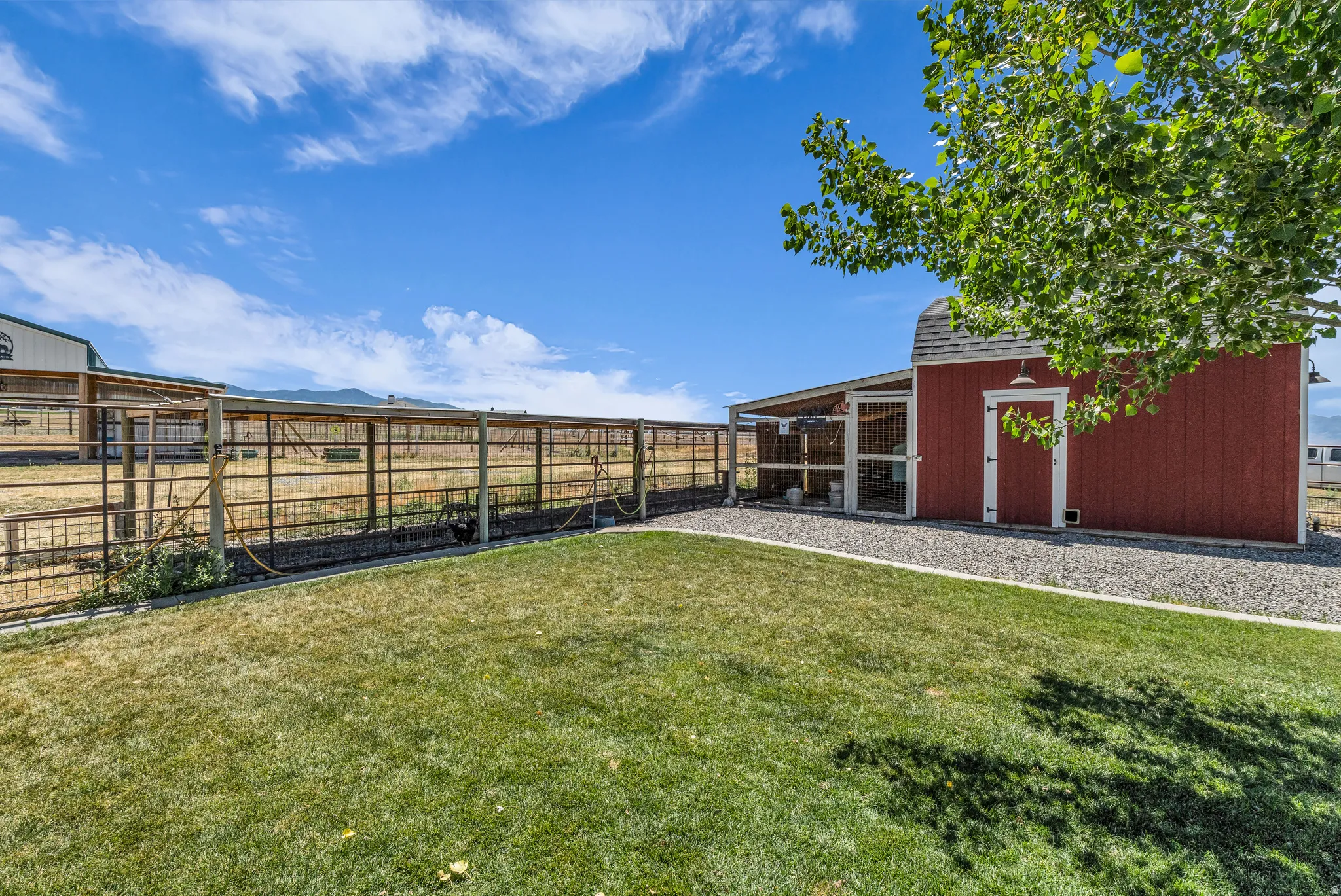 Expansive grassy area featuring a red barn structure with white trim, a gravel perimeter, an attached covered enclosure, and metal fencing