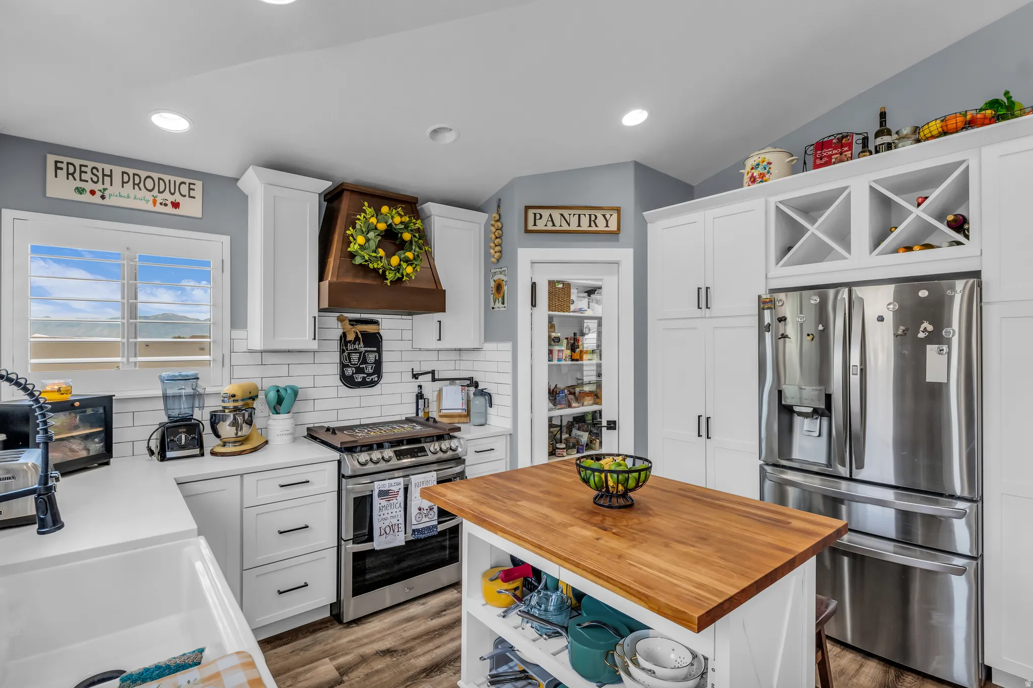 Bright kitchen featuring white shaker cabinetry, stainless steel appliances, a wood-finish range hood, white subway tile backsplash, and a central island with a butcher block countertop