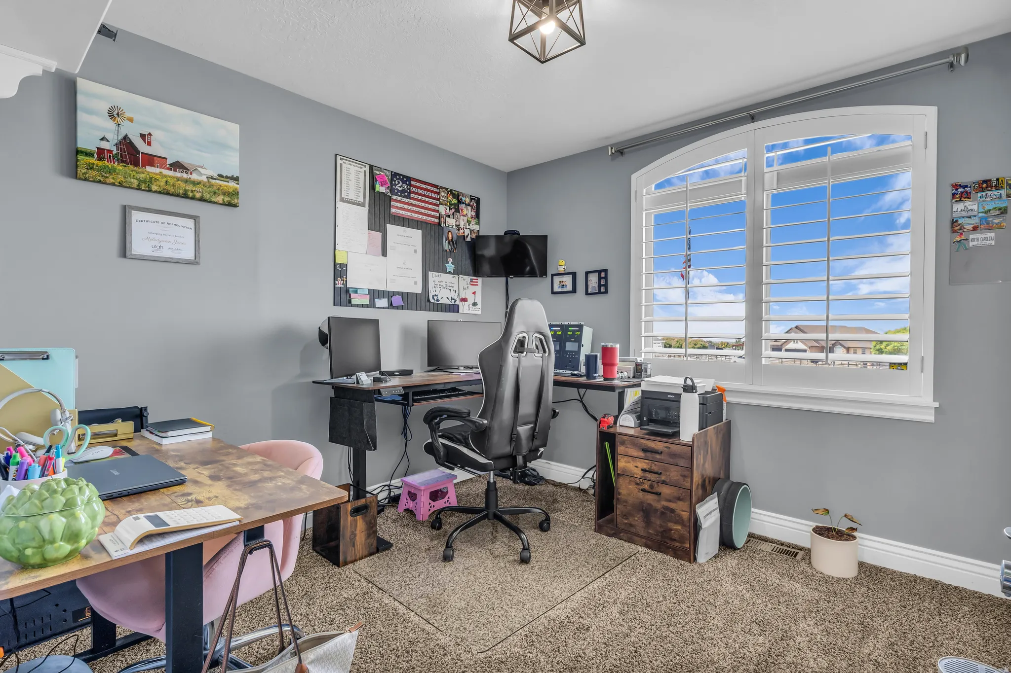 Bedroom currently used as an office with arched window with white plantation shutters, light gray walls, textured carpet flooring, and a decorative ceiling light fixture