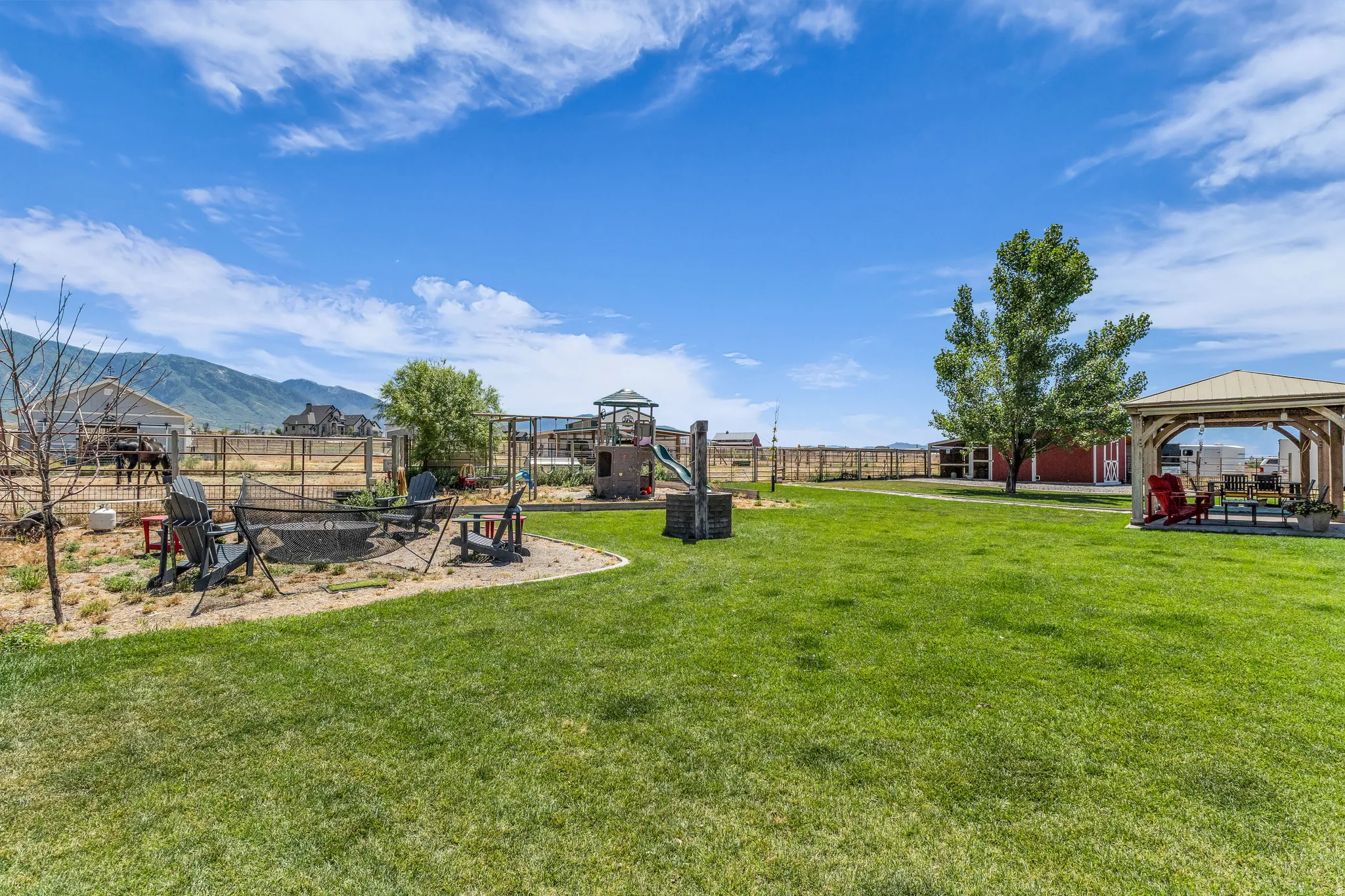 Expansive backyard featuring a large green lawn, a built-in fire pit area with seating, a wooden play structure, and a covered gazebo