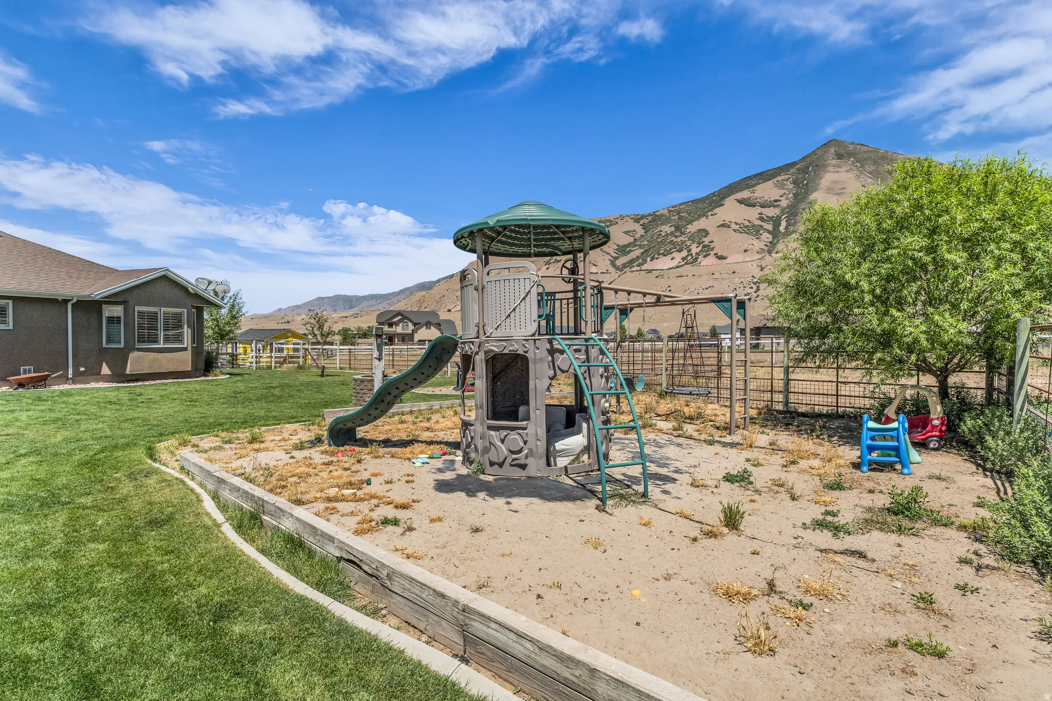 Spacious backyard featuring a large play structure, a sand-filled area, a fenced perimeter, and a distant mountainous backdrop