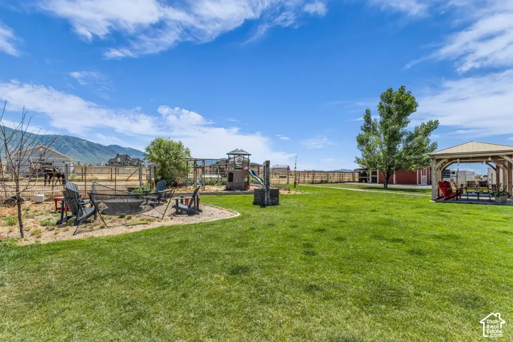 Expansive backyard featuring a large green lawn, a built-in fire pit area with seating, a wooden play structure, and a covered gazebo