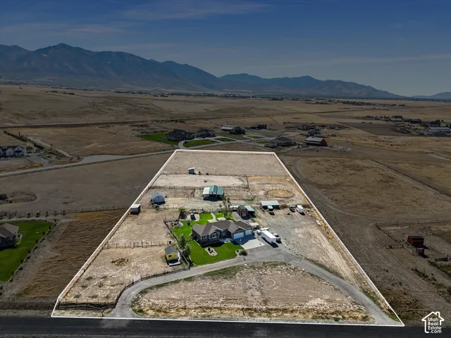 Expansive property with a single-story residence, asphalt driveway, multiple outbuildings, fenced perimeter, and mountainous backdrop