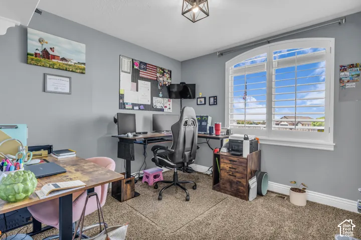 Bedroom 2 currently used as an office with arched window with white plantation shutters, light gray walls, textured carpet flooring, and a decorative ceiling light fixture