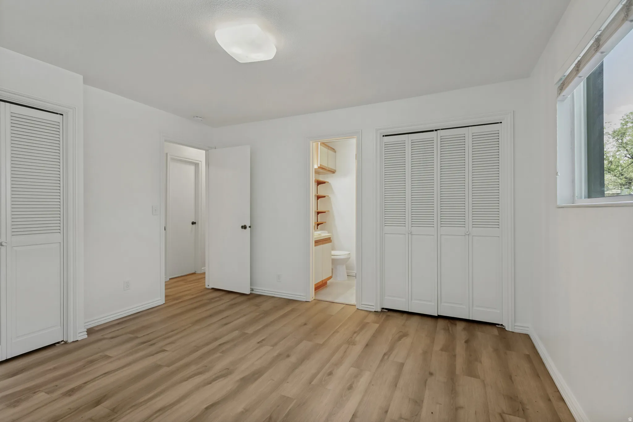 Wood-finish flooring, multiple louvered bifold closet doors, overhead lighting, and a window with natural light