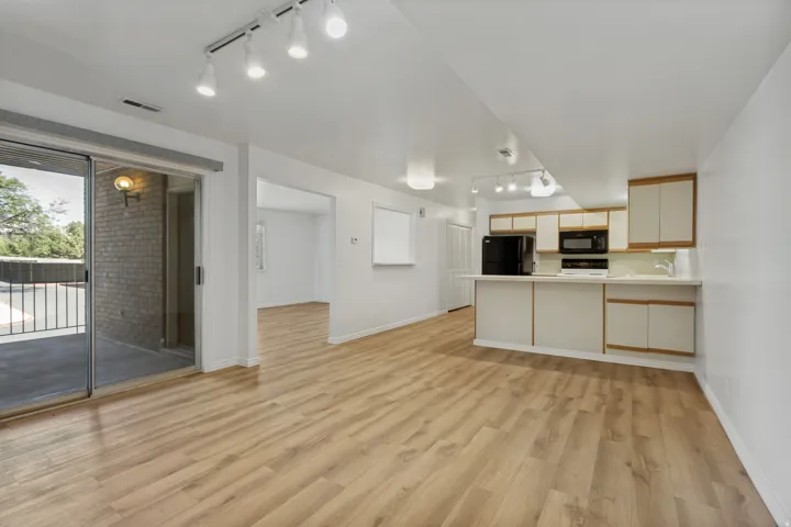 Wood-finish flooring throughout, kitchen with built-in cabinetry, black appliances, track lighting, and a sliding glass door to an outdoor patio