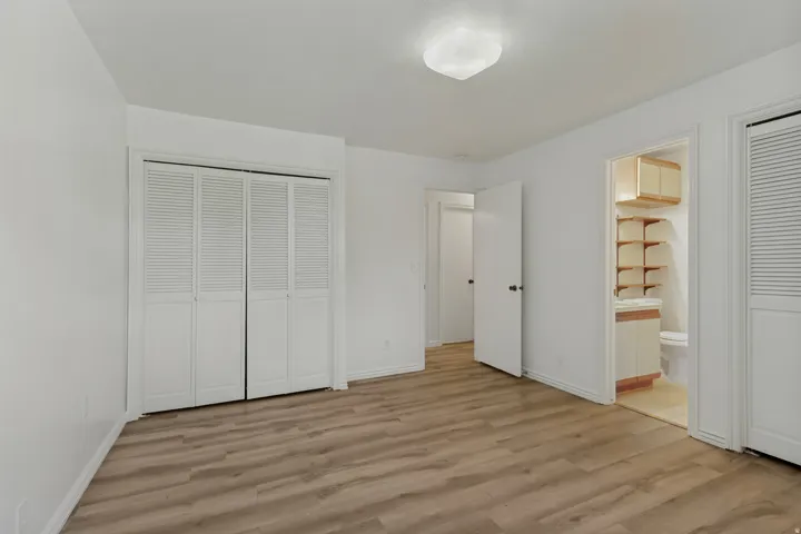 Wood-finish flooring, white walls, louvered bi-fold closet doors, flush-mount ceiling light, and an en-suite bathroom with built-in shelving