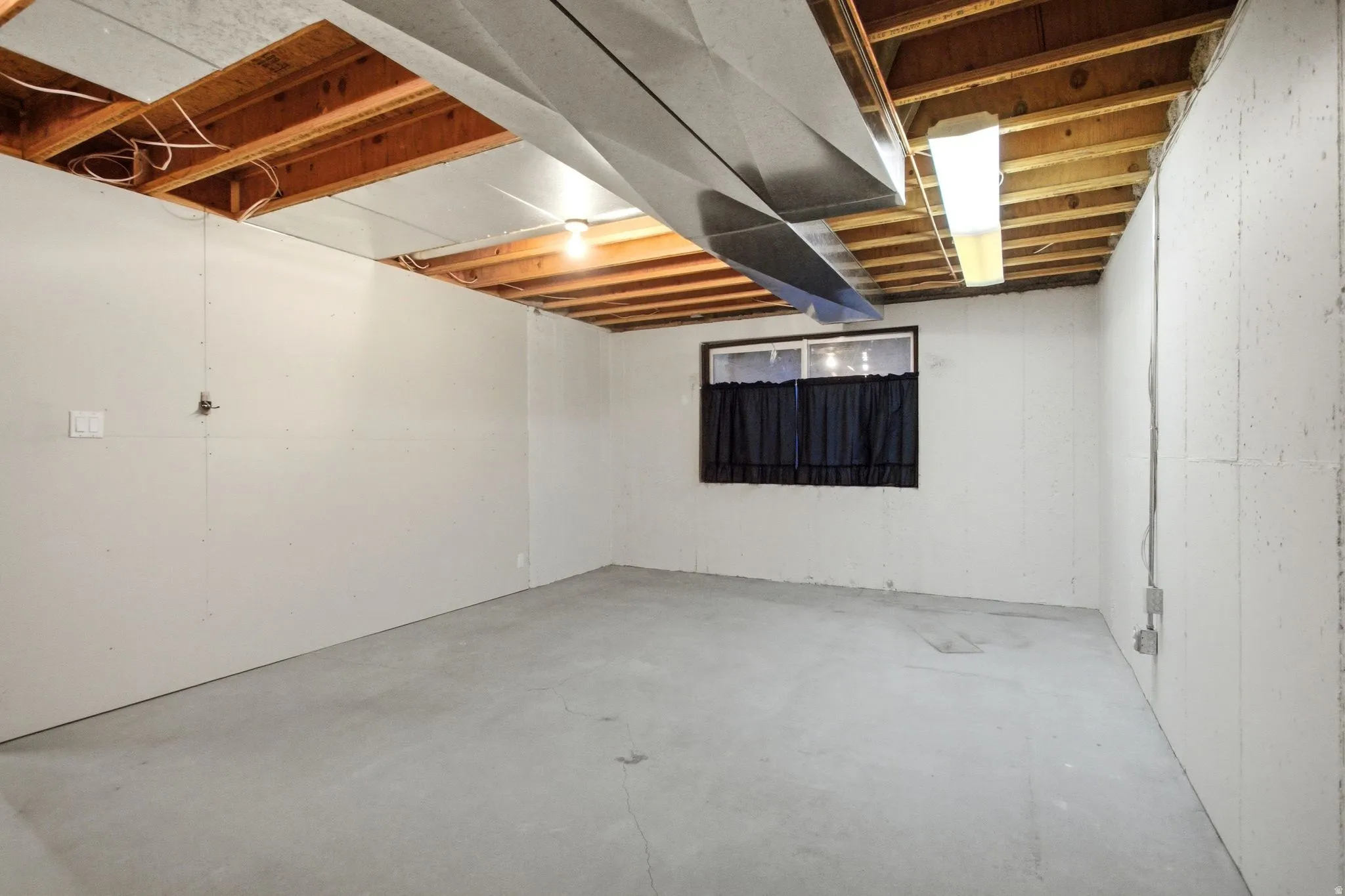 Unfinished basement space featuring exposed wooden ceiling joists, concrete flooring, and a single window