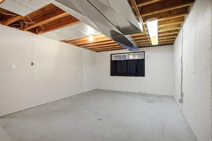 Unfinished basement space featuring exposed wooden ceiling joists, concrete flooring, and a single window
