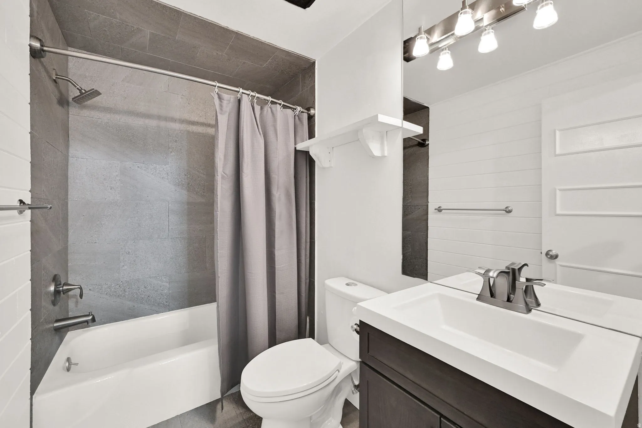 Contemporary bathroom featuring a white tub with gray tiled surround, a dark wood-finish vanity with an integrated white sink, a wall-mounted mirror, and brushed nickel fixtures