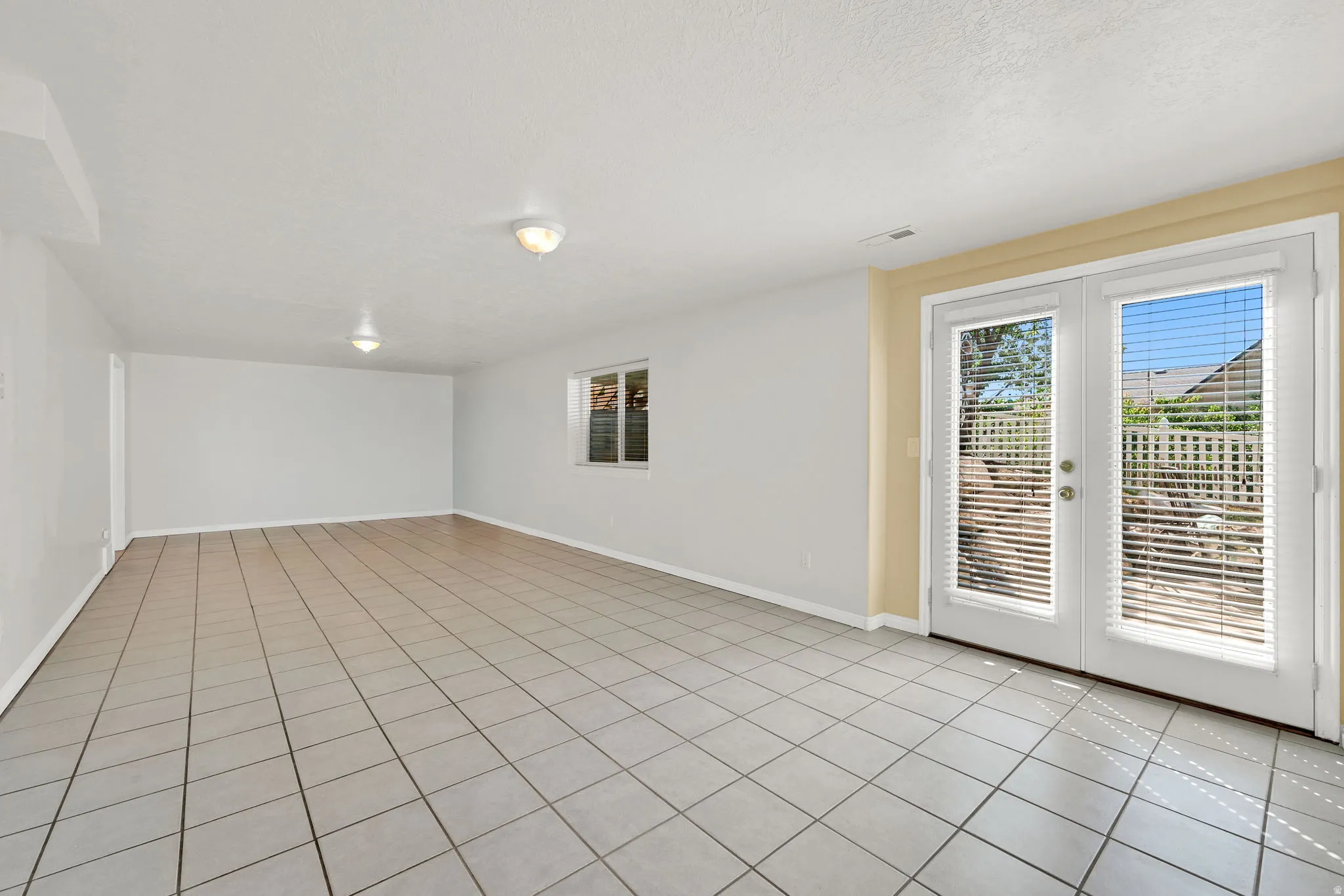 Spacious room featuring light-toned tile flooring, white walls, and a pair of white French doors with integrated blinds