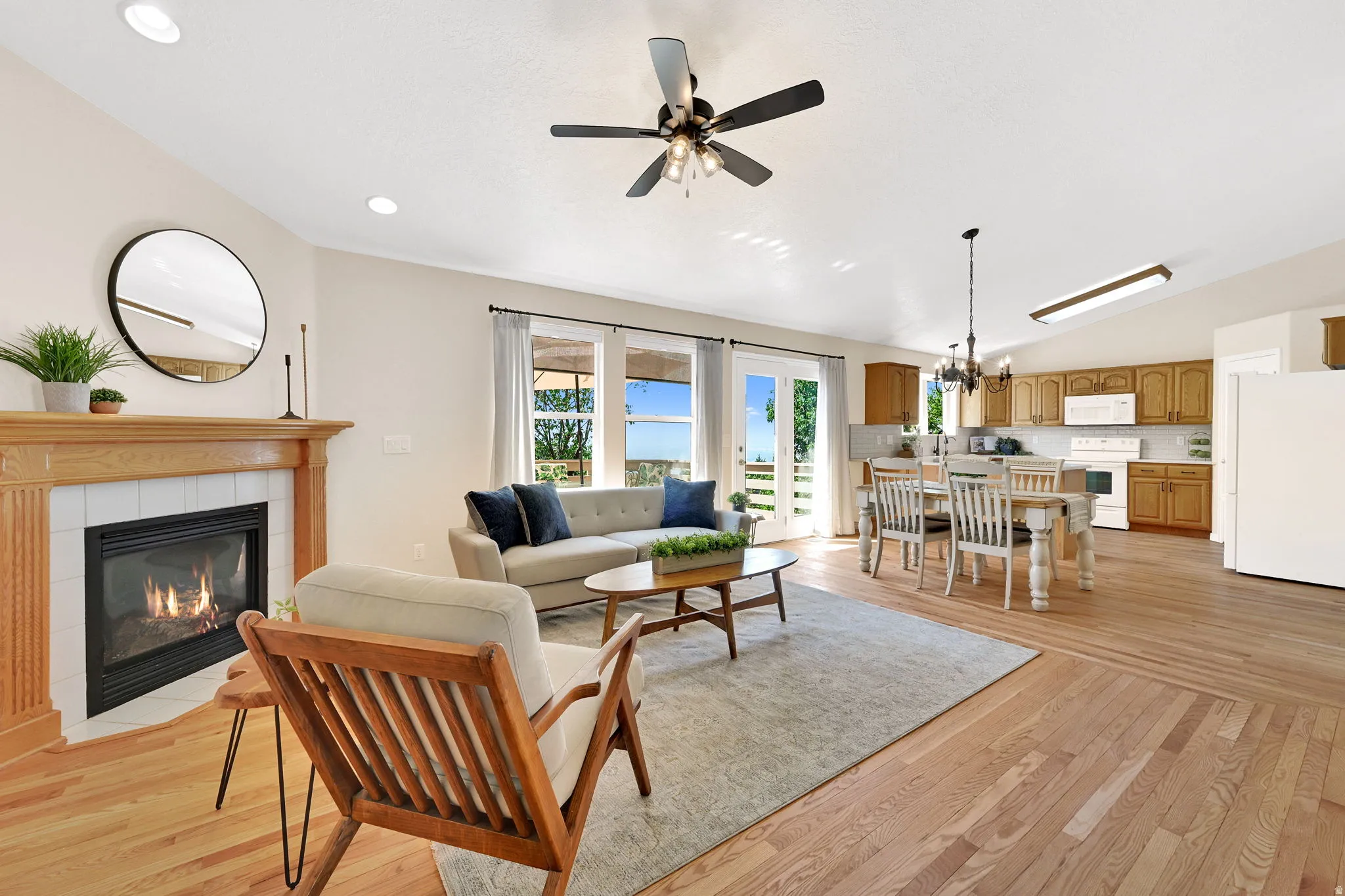 Open-concept living space featuring a fireplace with wood mantel and tile surround, wood-finish flooring, a kitchen with wood cabinetry and white appliances, and double glass doors opening to outdoor space