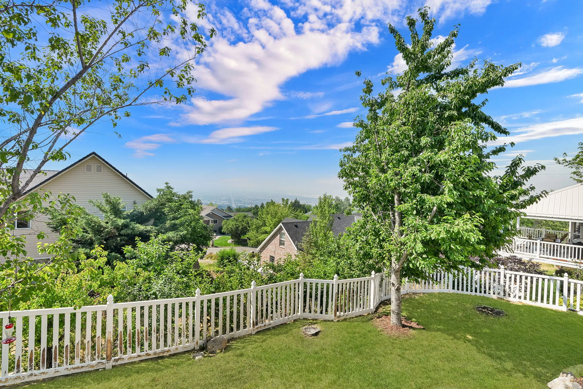Fenced backyard featuring a manicured lawn, established trees, and a distant cityscape