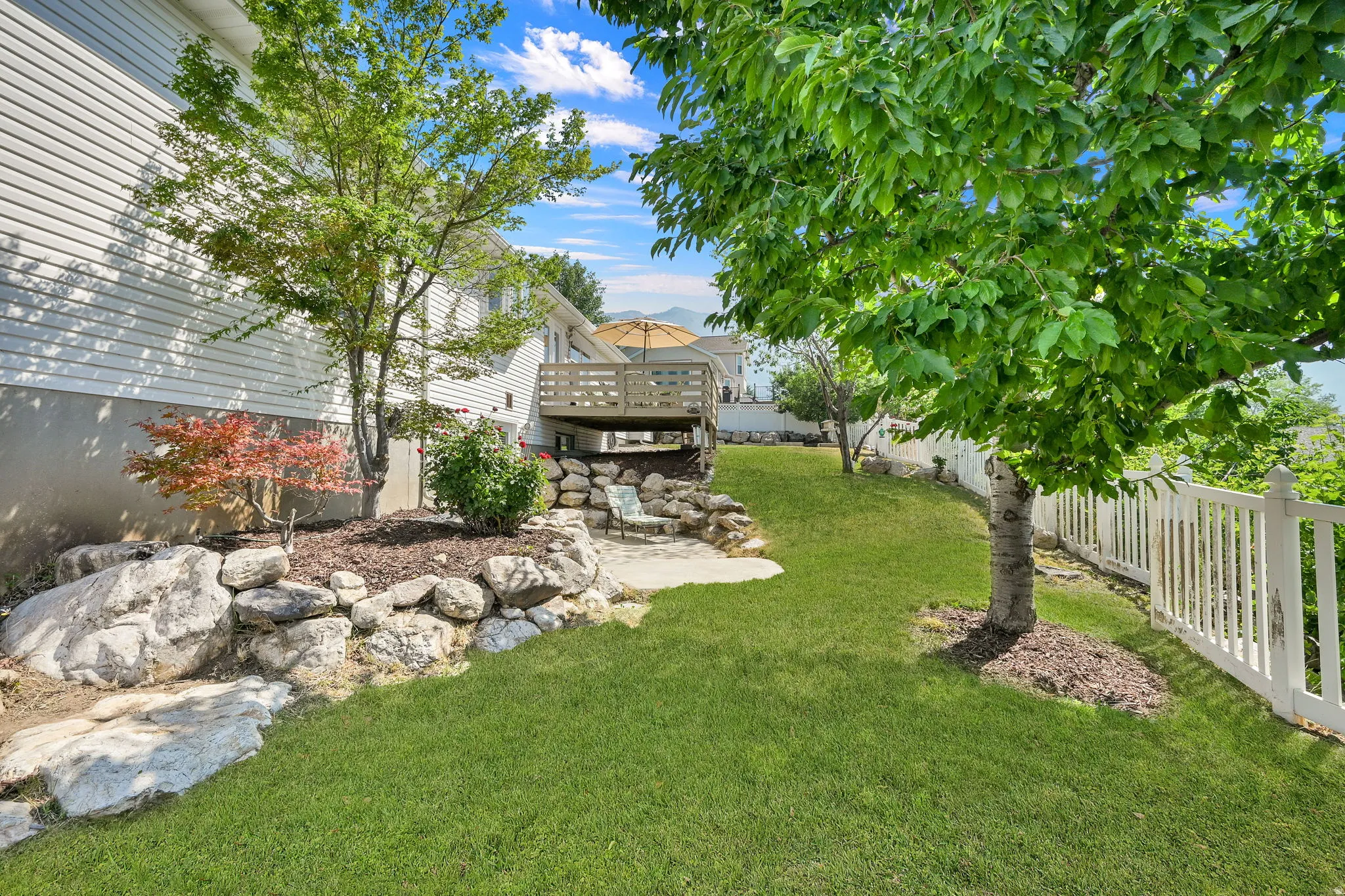 Expansive backyard featuring a manicured lawn, established trees, a white picket fence, a multi-level rock retaining wall, and a raised wood deck