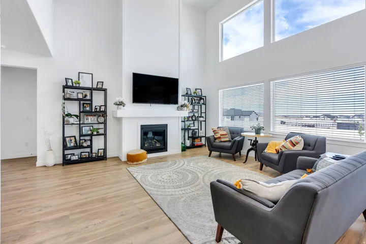 Spacious living area featuring high ceilings, large windows with blinds, a built-in fireplace with a white mantle, and light wood-finish flooring