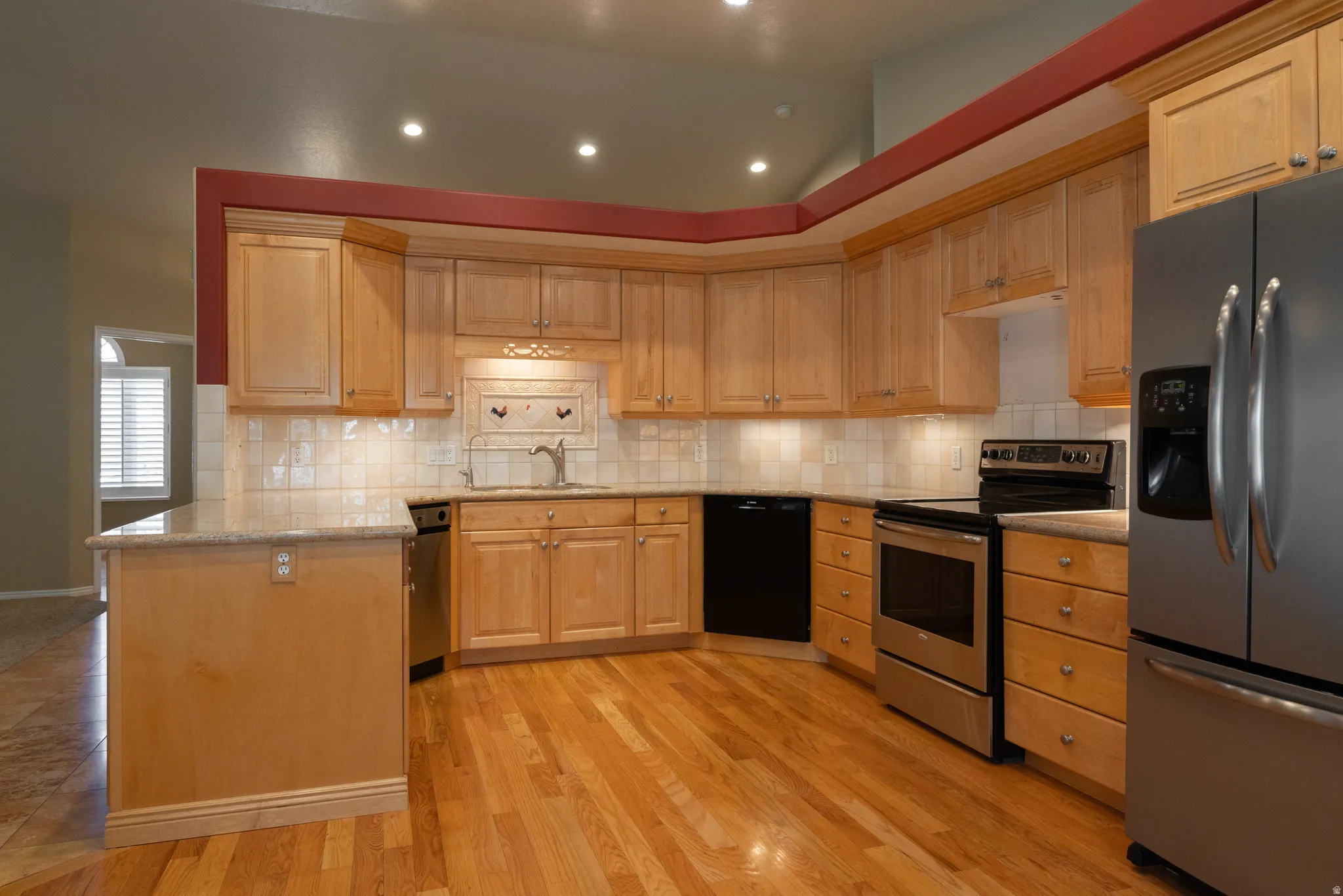 Open-concept kitchen featuring light wood cabinetry, stainless steel appliances, a white tile backsplash, and wood-finish flooring