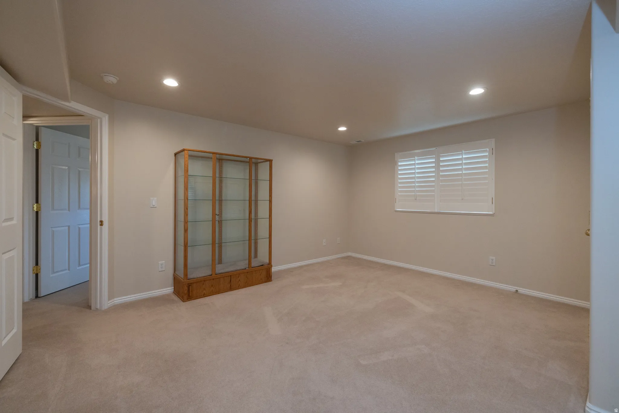 Spacious room with plush carpeting, recessed lighting, neutral wall tones, and a window featuring white plantation shutters