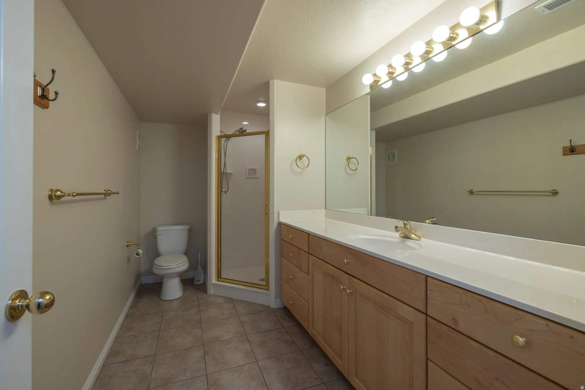 Bathroom featuring a gold-framed shower enclosure, wood-finish vanity with ample storage, a wide countertop, an expansive vanity mirror, and tile flooring