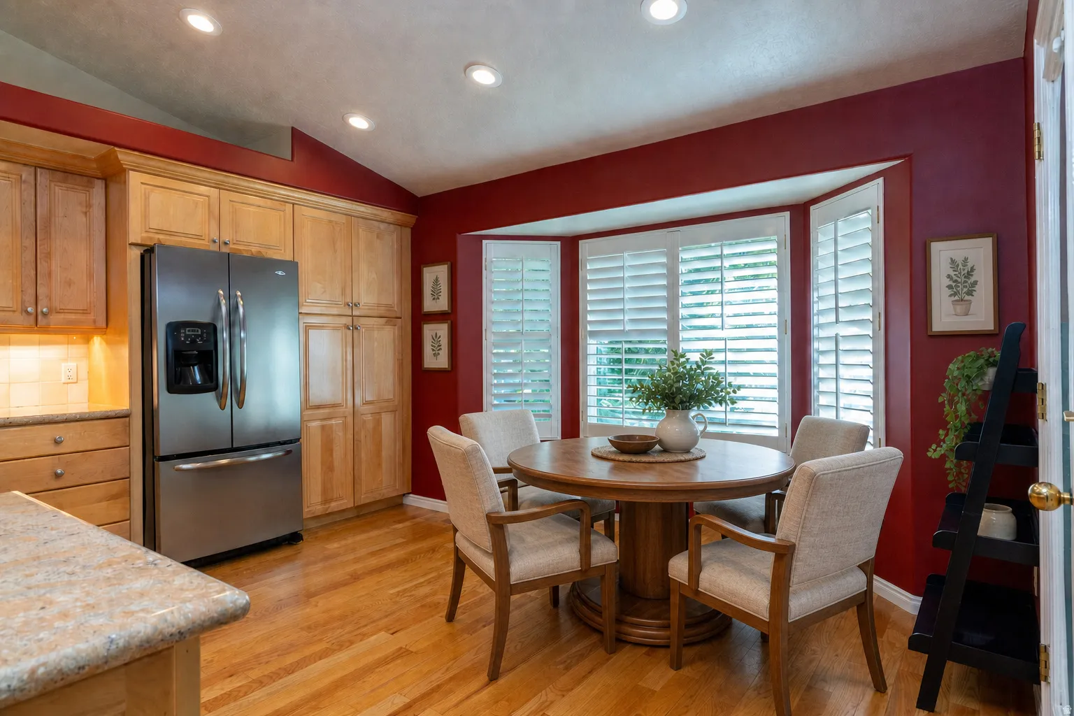 Virtually Staged - Kitchen dining area featuring wood-finish flooring, light wood cabinetry, a stainless steel refrigerator, and a bay window with white plantation shutters