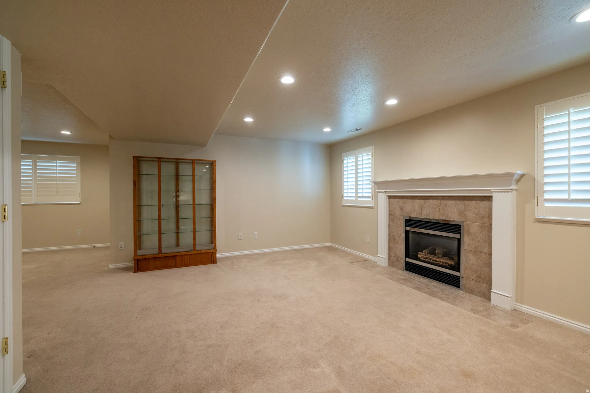 Spacious room featuring a fireplace with a tile surround and white mantel, recessed lighting, neutral carpeting, and two windows with white plantation shutters