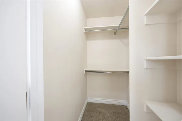 Walk-in closet featuring light-toned carpeting, white baseboards, multiple white shelving units, and chrome hanging rods