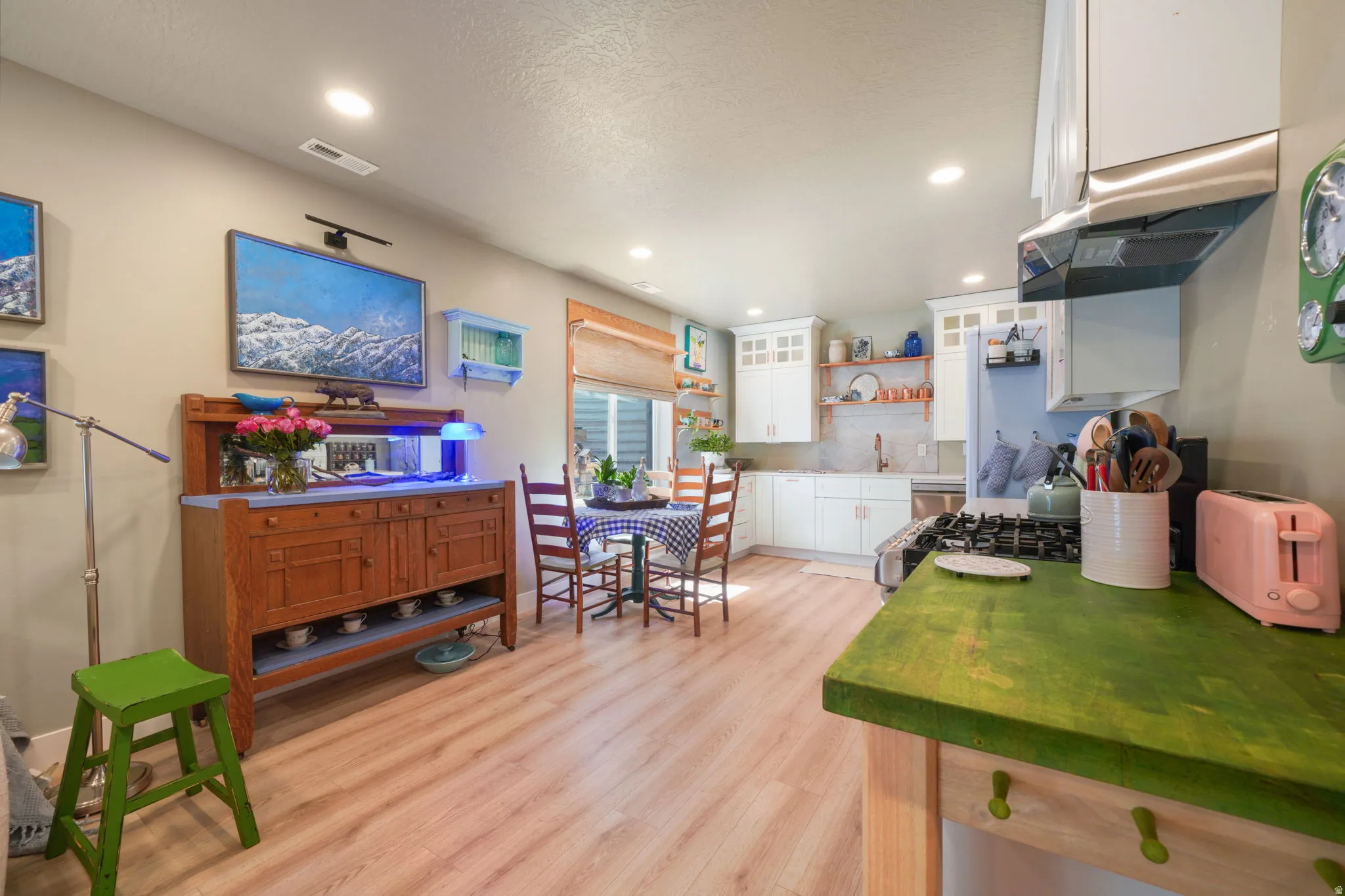 Open-concept kitchen and dining area featuring light wood-finish flooring, recessed lighting, white upper and lower cabinetry, a stainless steel range hood, and a built-in kitchen sink