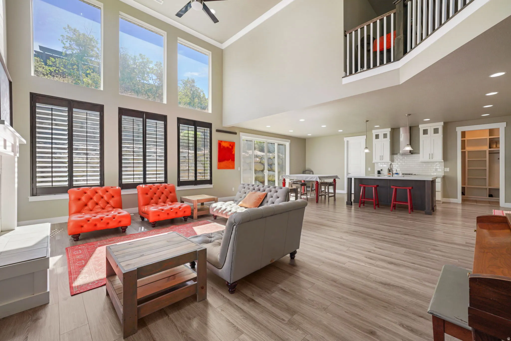 Two-story living area with high ceilings, wood-finish flooring, a white mantel, and a multi-level window arrangement featuring lower plantation shutters