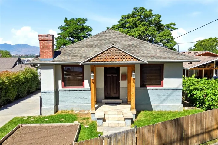 Gable roofline with asphalt shingles, red brick chimney, covered entry with wood columns, decorative wood shingle detailing, and a wooden fence