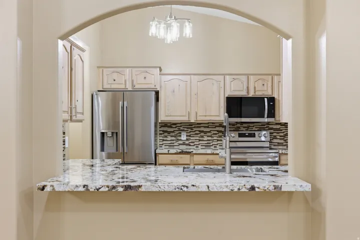 Kitchen pass-through featuring an arched entryway, stone-look countertop, stainless steel appliances, mosaic tile backsplash, and light-toned cabinetry