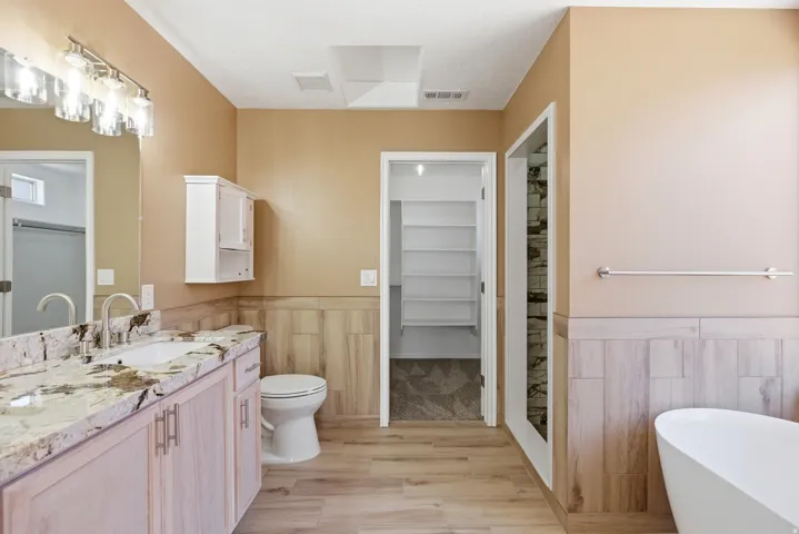 Bathroom featuring a double vanity with granite countertop, wood-finish flooring, a freestanding tub, and a separate shower with a stone-look tile surround