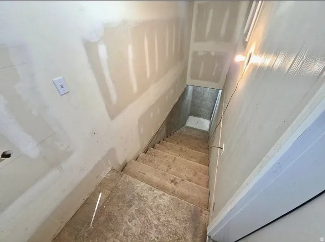 Unfinished lower-level staircase featuring exposed wood treads and risers, drywall-finished walls, and an overhead light fixture