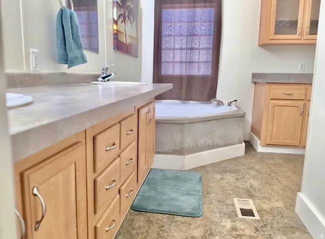 Bathroom featuring a built-in corner tub, light wood-finish cabinetry, a single vanity with an integrated sink, a tiled floor, and a window with privacy glass