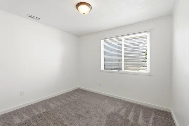 Light-filled room featuring a single window with horizontal blinds, neutral wall paint, gray textured carpet, white baseboards, and a semi-flush mount ceiling light fixture