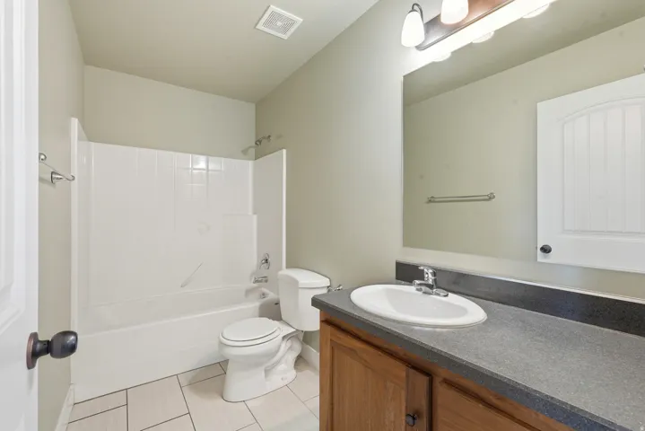Bathroom featuring a white tub and shower combination with a tile surround, a white toilet, wood-finish vanity with a dark countertop and integrated sink, a large wall-mounted mirror, and tile flooring