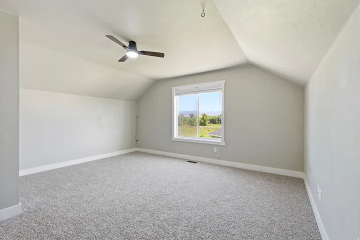 Carpeted room with a vaulted ceiling, a large window, a ceiling fan with integrated lighting, and white baseboards
