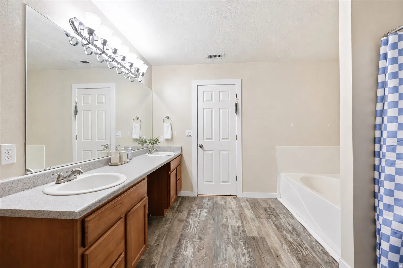 Bathroom featuring dual undermount sinks with chrome fixtures, a spacious vanity with wood-finish cabinetry, a large wall-mounted mirror, and wood-finish flooring