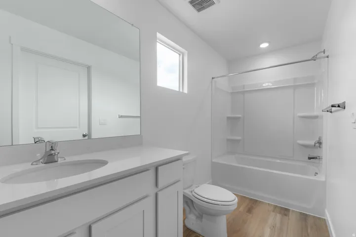 Bathroom featuring a vanity with an integrated sink and light-toned countertop, a large mirror, a toilet, a bathtub with a shower surround, and wood-finish flooring