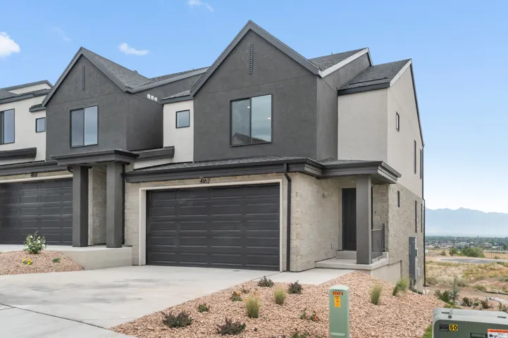 Modern townhome exterior featuring a two-car garage with dark gray door, multi-toned stucco and stone facade, dark gray window frames, and a covered entry
