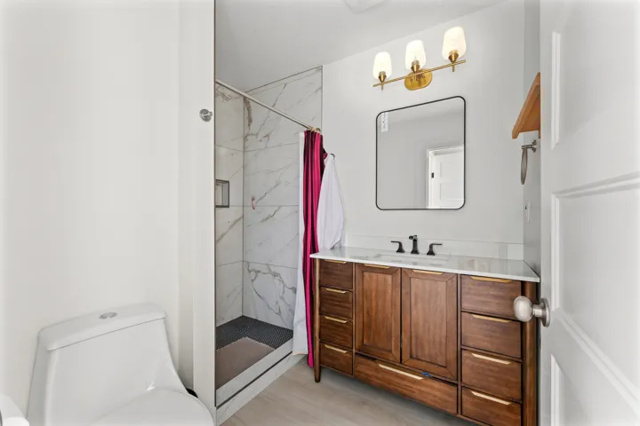 Bathroom featuring a glass-enclosed shower with white and gray tiled walls, a wood-finish vanity with a white countertop and integrated sink, a rectangular mirror with a black frame, and a three-light vanity fixture with brass accents