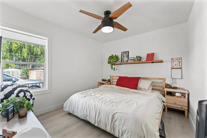 Bright room featuring a large window with white trim, wood-finish flooring, a contemporary ceiling fan with dark accents, a built-in wood-toned floating shelf, and white walls