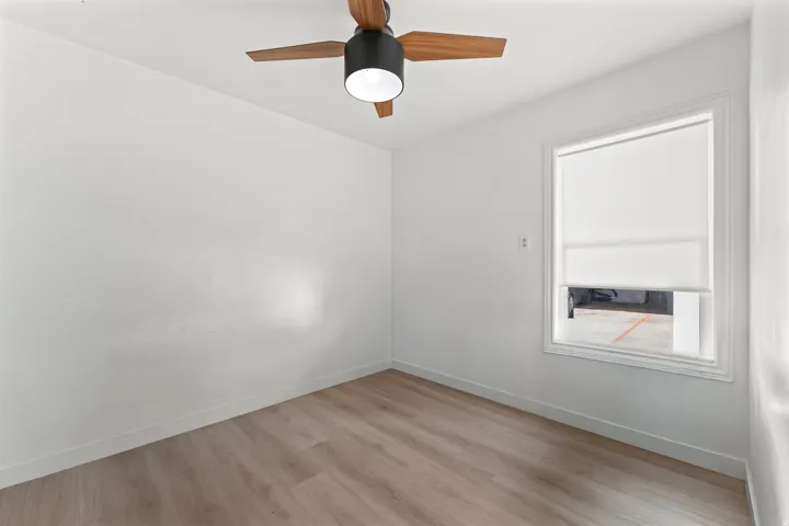 Bright interior space featuring a ceiling fan with wood-finish blades, a white roller blind on a window, wood-finish flooring, and white baseboards