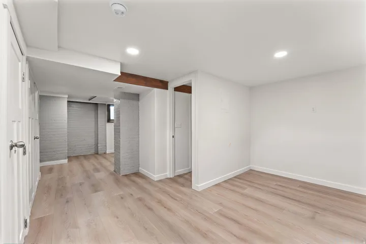 Finished interior space featuring light wood-finish flooring, white painted walls, recessed lighting, white baseboards, and a visible wood beam accent