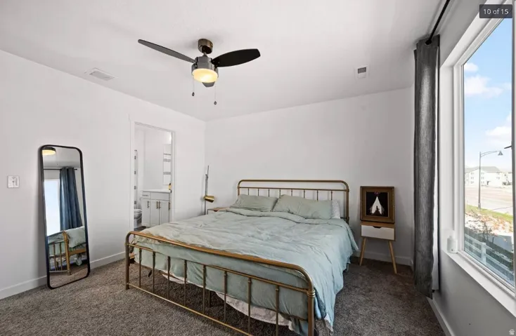 Carpeted room featuring a ceiling fan with integrated lighting, a large window with privacy curtains, and a doorway opening to a bathroom with a white vanity