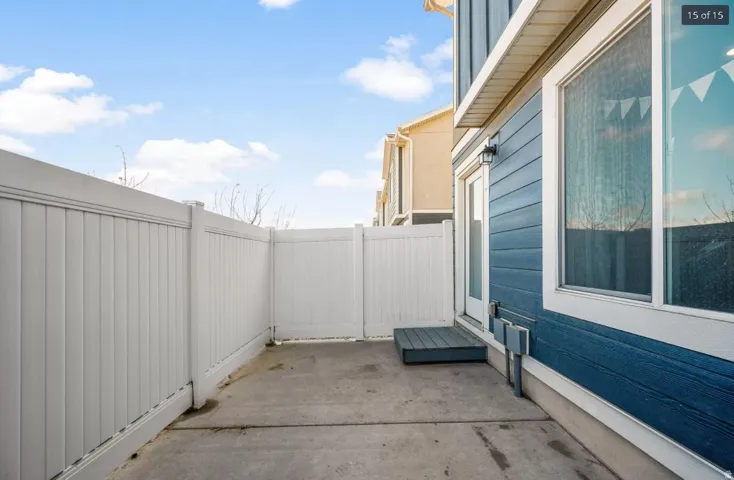 Concrete patio area featuring a white privacy fence, blue horizontal siding, white trim windows, and a rear entry door with an exterior light fixture