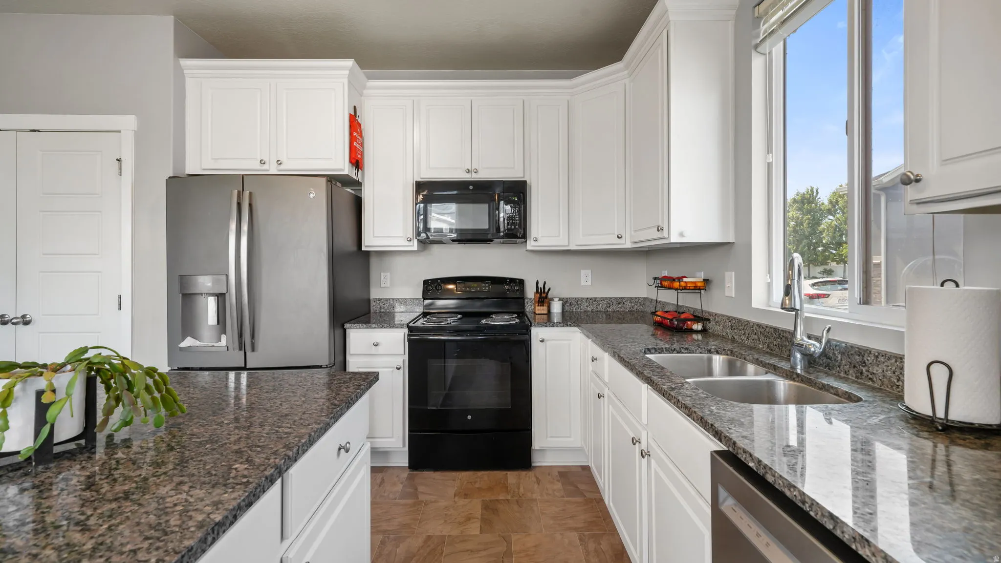 Kitchen featuring white cabinetry, dark stone countertops, stainless steel refrigerator, black range, and dual basin sink with window
