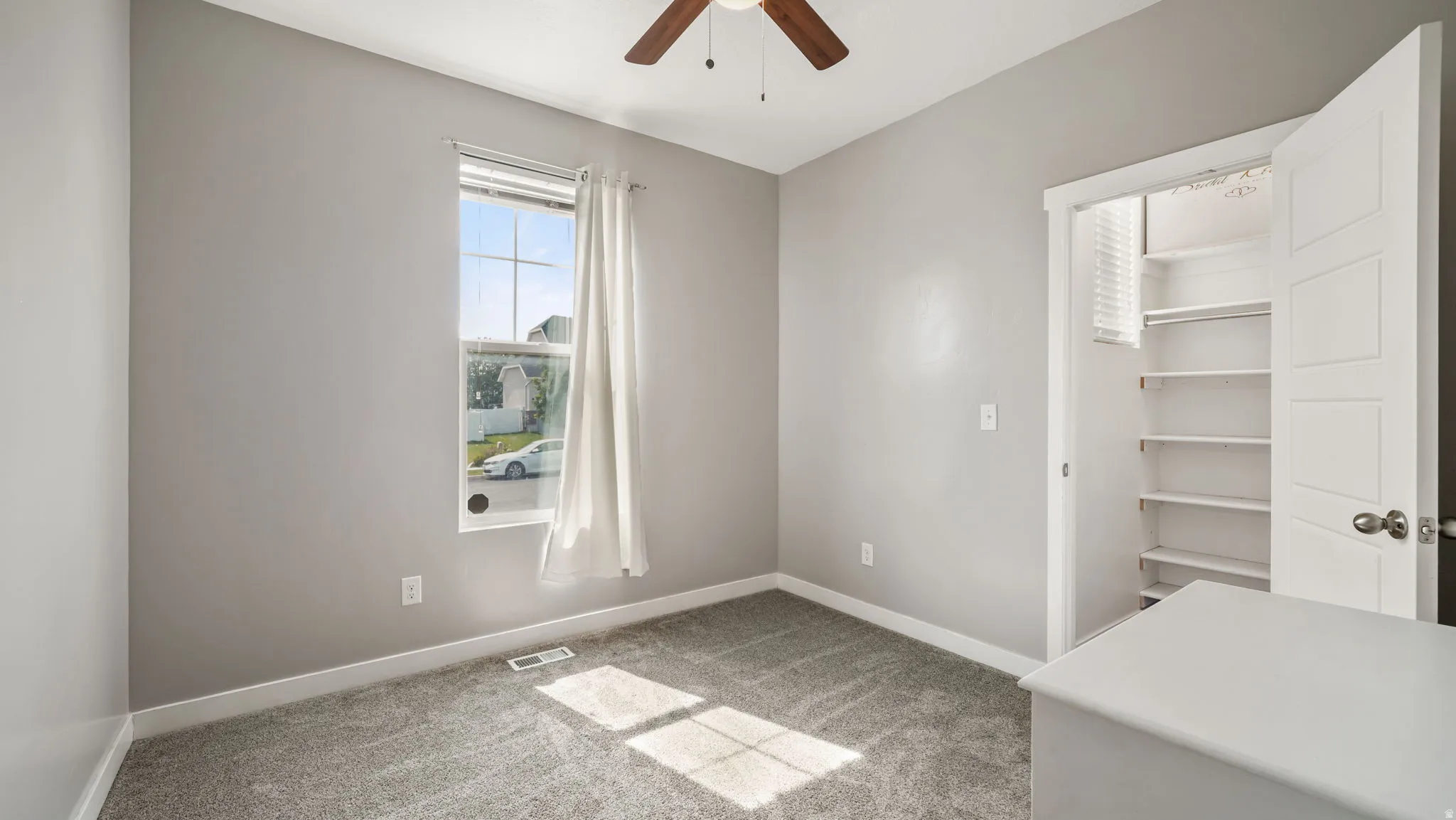 Neutral-toned room featuring a window with white blinds and drapes, a ceiling fan, plush grey carpeting, and a built-in closet with shelving